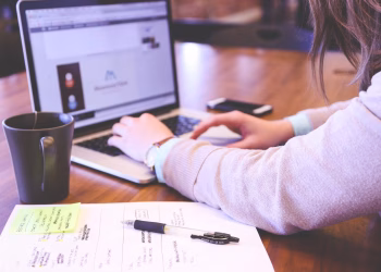Businesswoman typing on laptop with notes and coffee on wooden desk.