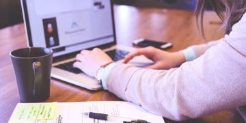 Businesswoman typing on laptop with notes and coffee on wooden desk.