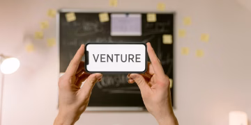 A smartphone displaying 'Venture' held by hands in an office setup. Ideal for business and startup themes.