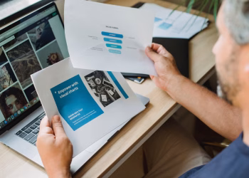 Crop anonymous male with beard looking at documents with graphic charts while sitting at table with open laptop