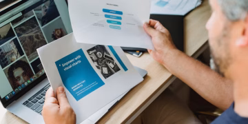 Crop anonymous male with beard looking at documents with graphic charts while sitting at table with open laptop