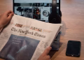 Close-up of hands holding a newspaper by a laptop and smartphone on a table.