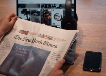 Close-up of hands holding a newspaper by a laptop and smartphone on a table.