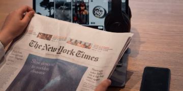 Close-up of hands holding a newspaper by a laptop and smartphone on a table.