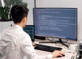 Adult male programmer coding on dual monitors in a modern indoor workspace.