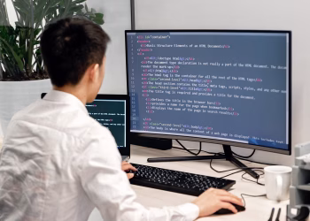 Adult male programmer coding on dual monitors in a modern indoor workspace.