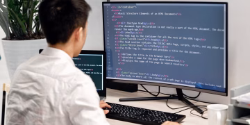 Adult male programmer coding on dual monitors in a modern indoor workspace.