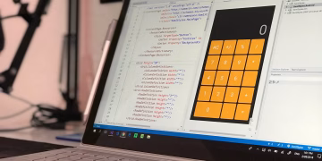 A laptop screen shows a coding application with a calculator design in a tech office setting.