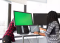 Two women working on laptops and monitors in a bright office setting, focused on technology and teamwork.