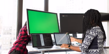 Two women working on laptops and monitors in a bright office setting, focused on technology and teamwork.