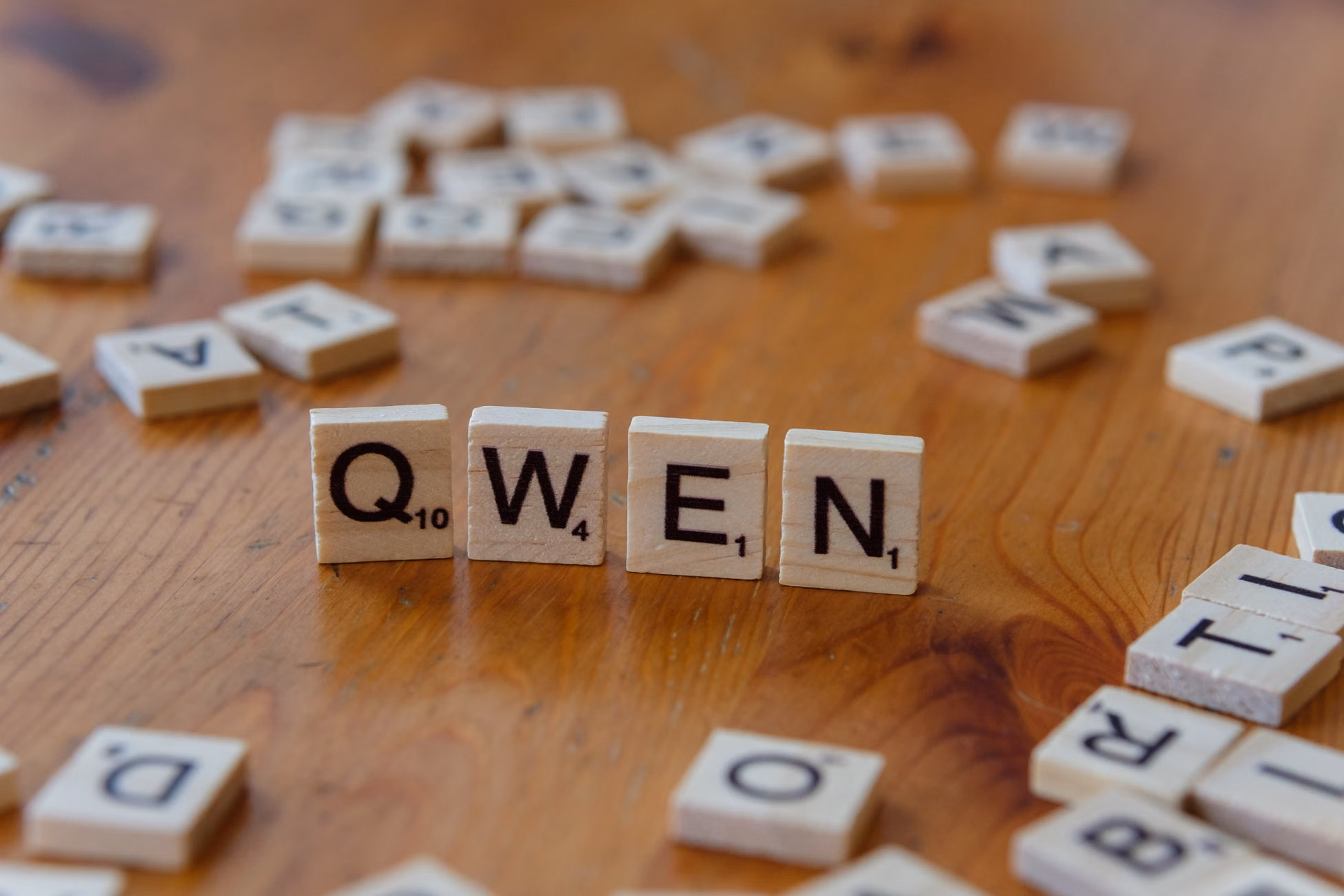 Wooden Scrabble tiles form the word 'QWEN' on a wooden surface, with scattered tiles in the background.