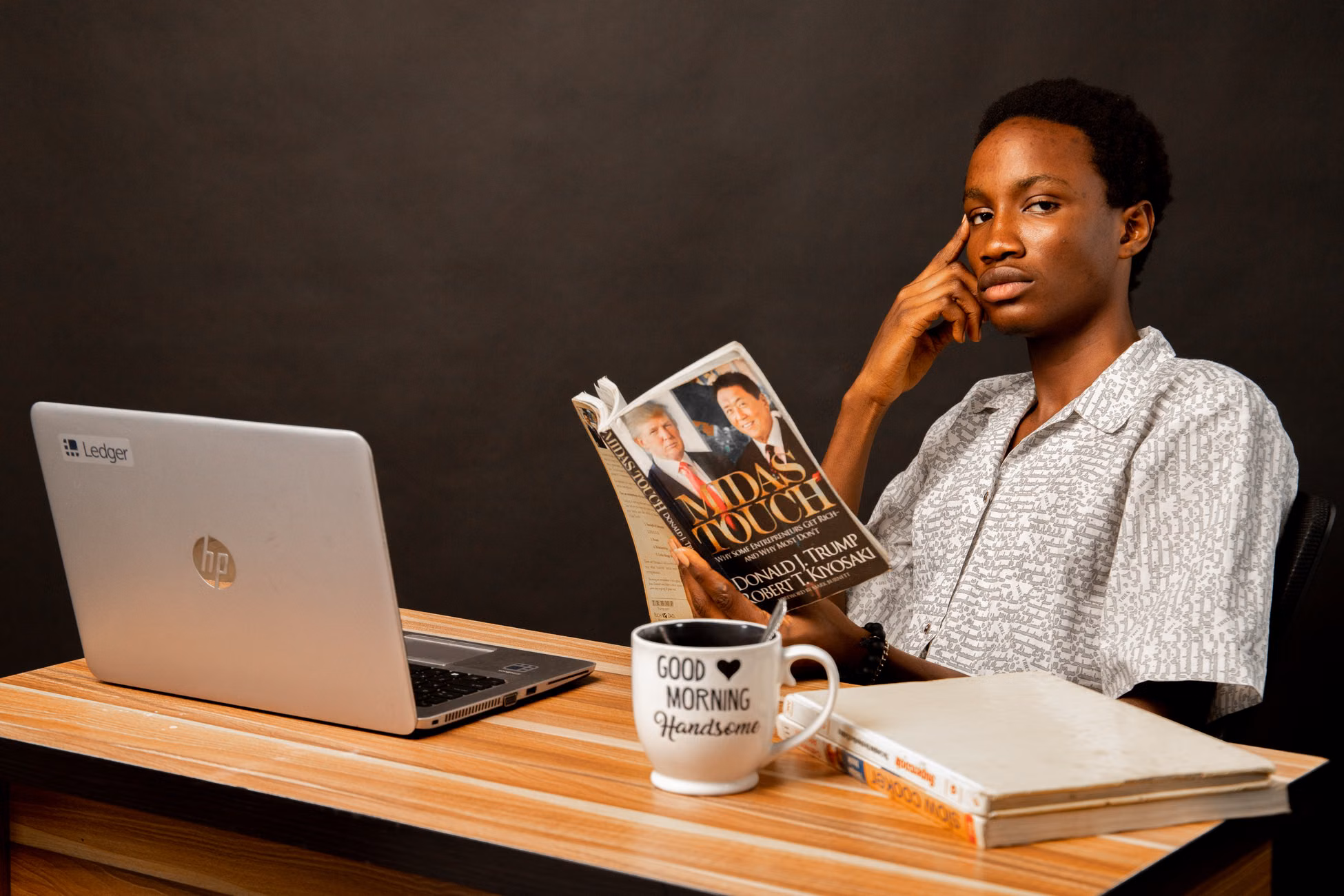 Young man reading a book at his desk with a laptop and a cup, embodying a relaxed study atmosphere.