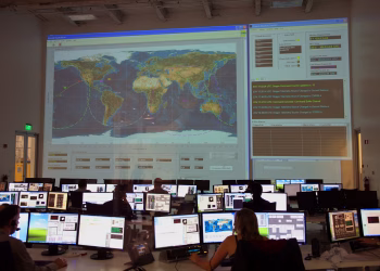 Back view of unrecognizable employees working on computers in flight control room with big interactive map on wall and analyzing data