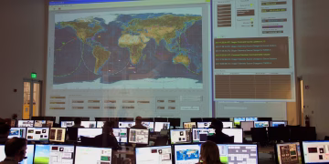 Back view of unrecognizable employees working on computers in flight control room with big interactive map on wall and analyzing data