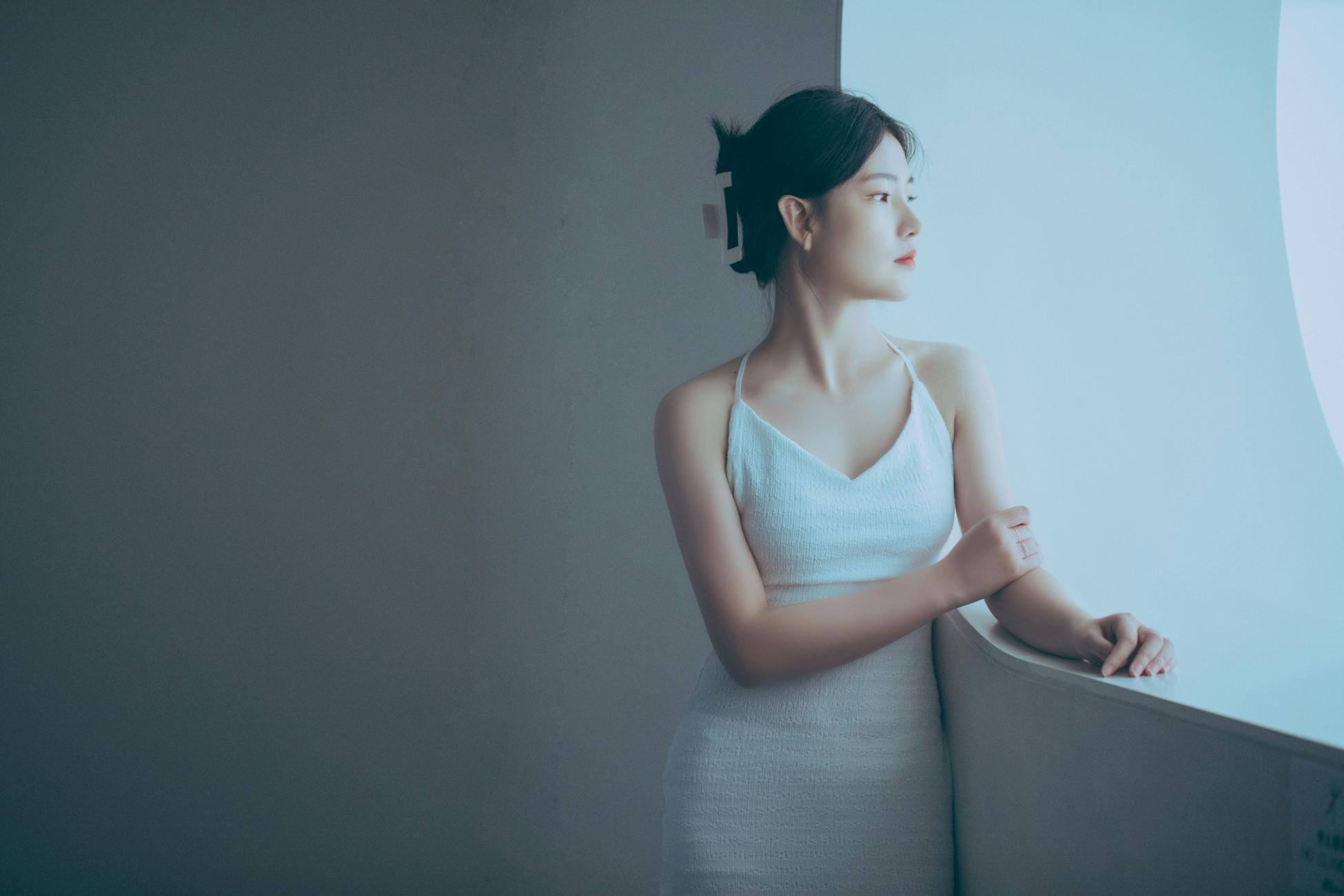 Stylish woman in a white dress gazing thoughtfully through a window indoors.