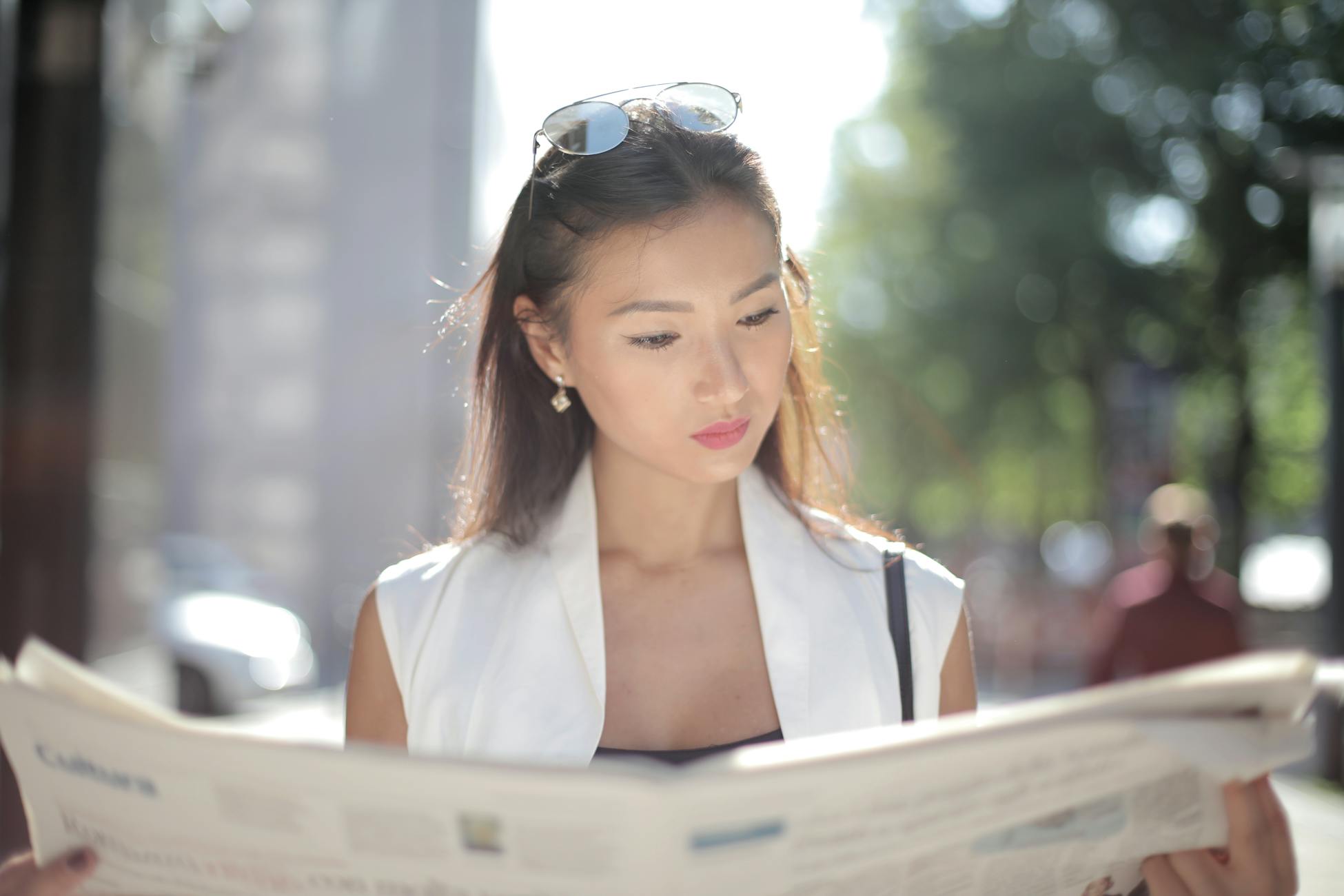 Elegant woman with sunglasses reads a newspaper on a sunny day outside.