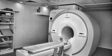 Black and white image of a modern MRI machine in a hospital room with overhead ceiling art.