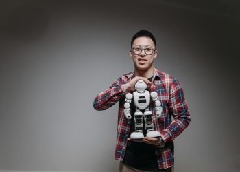 Asian man with eyeglasses holding a toy robot in a studio with a gray background.