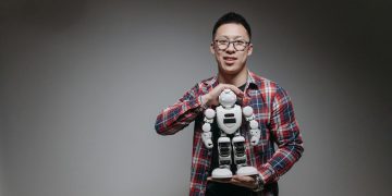 Asian man with eyeglasses holding a toy robot in a studio with a gray background.