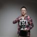 Asian man with eyeglasses holding a toy robot in a studio with a gray background.
