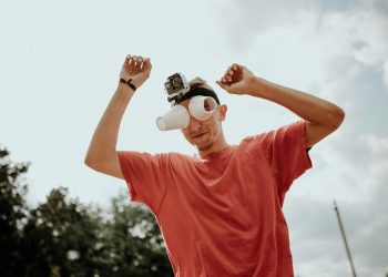 A man outdoors in a red t-shirt with a GoPro and makeshift binoculars made from cups, arms raised playfully.