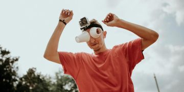 A man outdoors in a red t-shirt with a GoPro and makeshift binoculars made from cups, arms raised playfully.