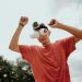A man outdoors in a red t-shirt with a GoPro and makeshift binoculars made from cups, arms raised playfully.