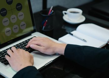 Close-up of hands typing on a laptop displaying an organizational chart, a notebook, and coffee nearby.