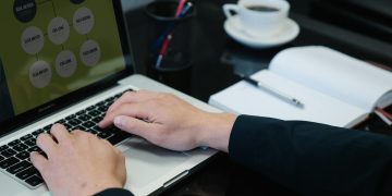 Close-up of hands typing on a laptop displaying an organizational chart, a notebook, and coffee nearby.