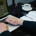 Close-up of hands typing on a laptop displaying an organizational chart, a notebook, and coffee nearby.