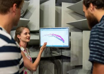 Three engineers discussing sound waves displayed on a monitor in a professional sound studio.