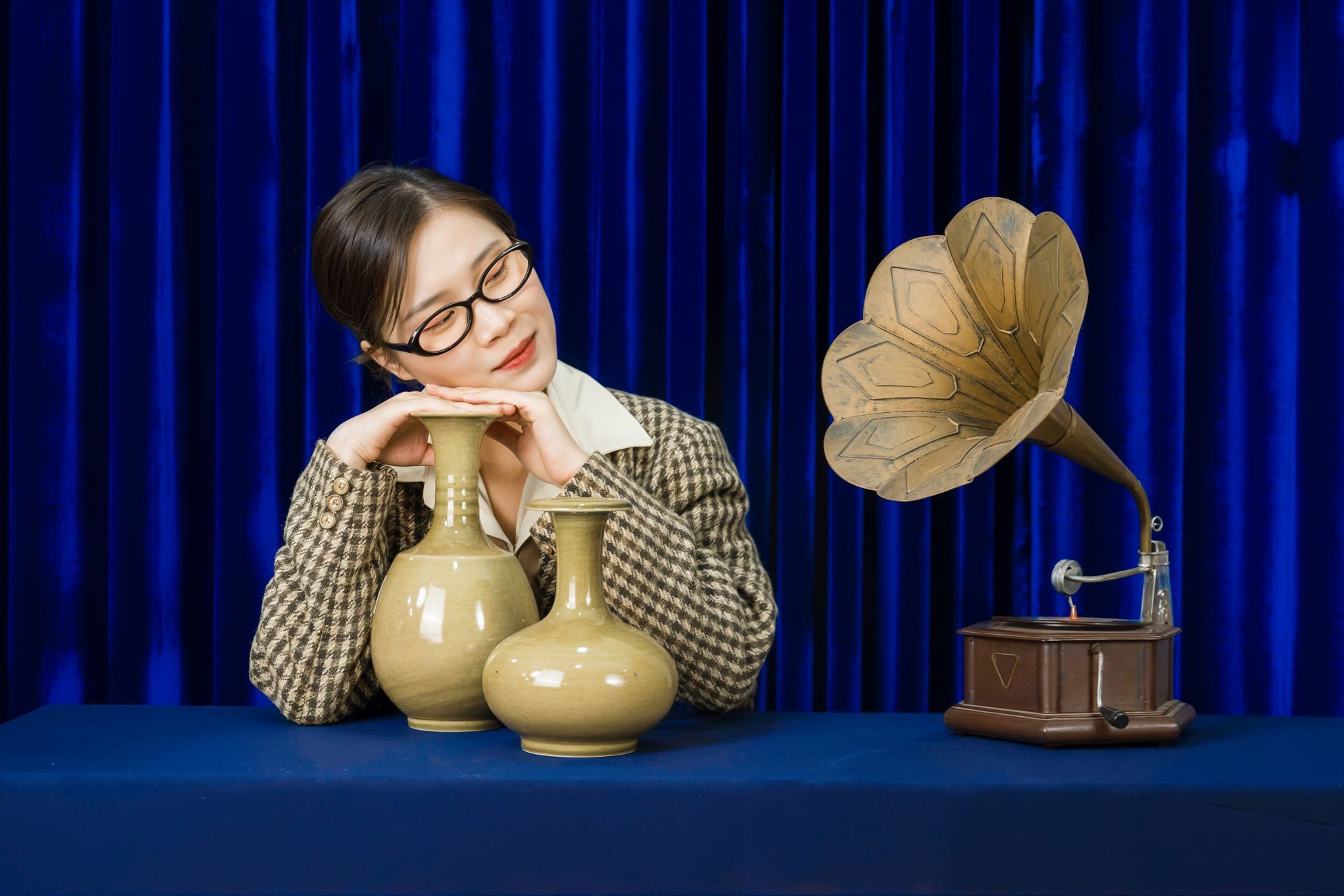 A woman in glasses poses with vintage vases and a gramophone against a blue curtain, evoking a classic style.