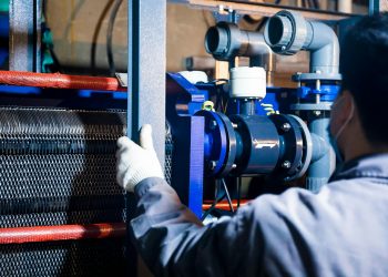A worker in blue sleeves inspects machinery pipes and equipment at an industrial plant.