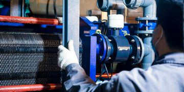 A worker in blue sleeves inspects machinery pipes and equipment at an industrial plant.