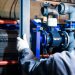 A worker in blue sleeves inspects machinery pipes and equipment at an industrial plant.