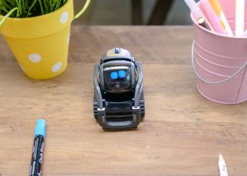 A small modern robot placed on a wooden desk surrounded by office supplies, showcases innovative technology.
