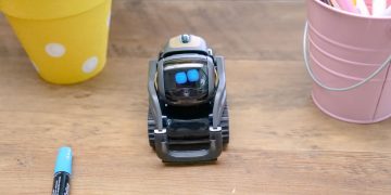 A small modern robot placed on a wooden desk surrounded by office supplies, showcases innovative technology.
