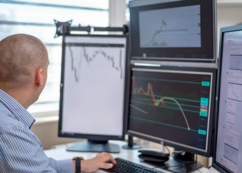 Trader analyzing financial data on multiple monitors in an office setting.