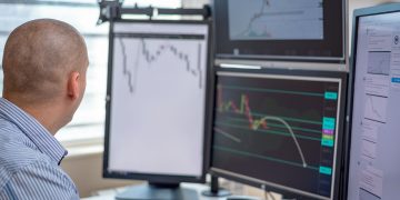 Trader analyzing financial data on multiple monitors in an office setting.