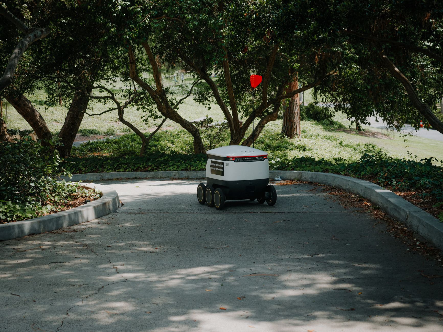 An autonomous delivery robot moves through a shaded pathway in a park, exemplifying modern logistics.