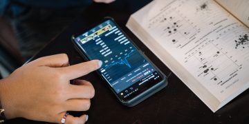 Hand pointing at smartphone displaying financial data, with open book nearby.