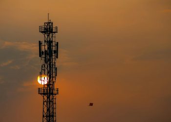 Silhouette of a telecommunication tower against a vibrant sunset sky with a distant kite.