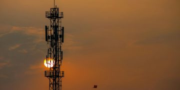 Silhouette of a telecommunication tower against a vibrant sunset sky with a distant kite.