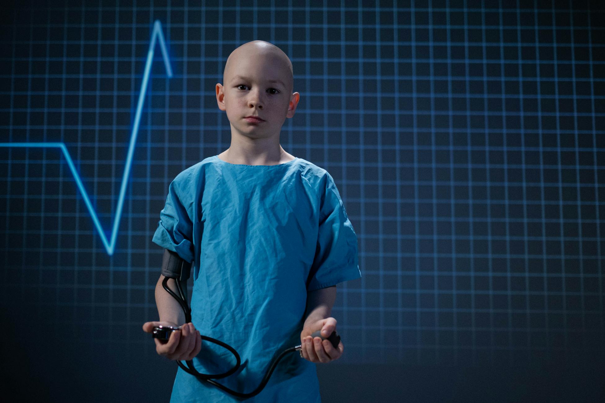 Bald child in hospital gown holding blood pressure monitor, standing before EKG grid.