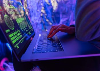 Close-up of hands typing on a laptop displaying cybersecurity graphics, illuminated by purple light.