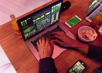 Hands typing on a laptop with coding, phone on desk, symbolizing cybersecurity.