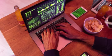 Hands typing on a laptop with coding, phone on desk, symbolizing cybersecurity.