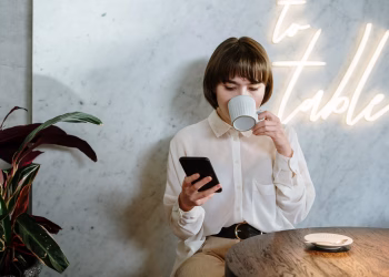 Stylish café scene with young woman enjoying her coffee, using a mobile phone under neon light ambiance.