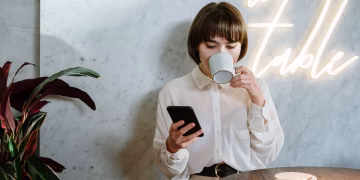 Stylish café scene with young woman enjoying her coffee, using a mobile phone under neon light ambiance.
