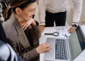 Businesswoman using laptop at office desk surrounded by colleagues, collaborating on project.